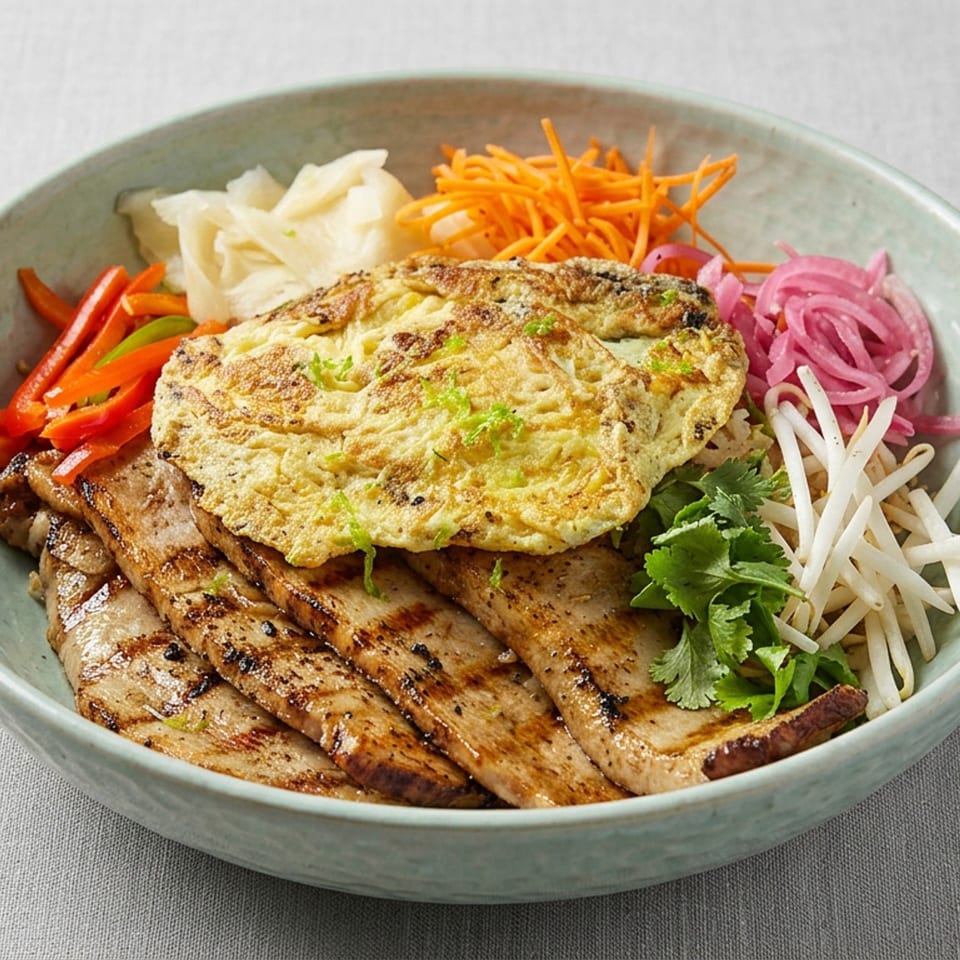 Bowl of grilled pork topped with a savory omelet, served with pickled vegetables, carrots, peppers, bean sprouts, and cilantro.