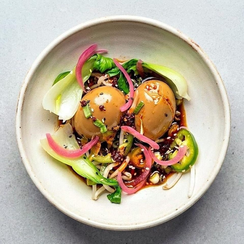 Bowl of jammy eggs with bok choy, noodles, pickled onions, peppers, herbs, and spicy sauce.